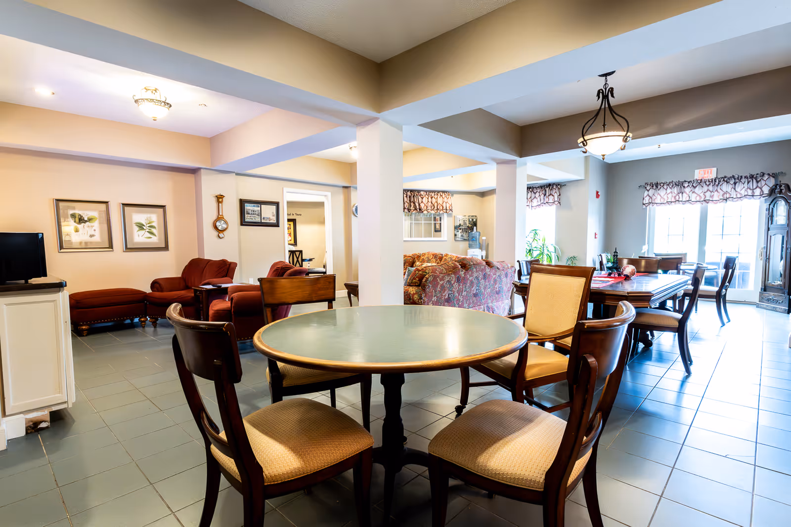 A bright and spacious common area in a senior living facility featuring a round table with four chairs in the foreground, a larger dining table with chairs in the background near windows with patterned valances, and a seating area with upholstered chairs and a sofa. The room has tiled floors, beige walls, and ceiling light fixtures.