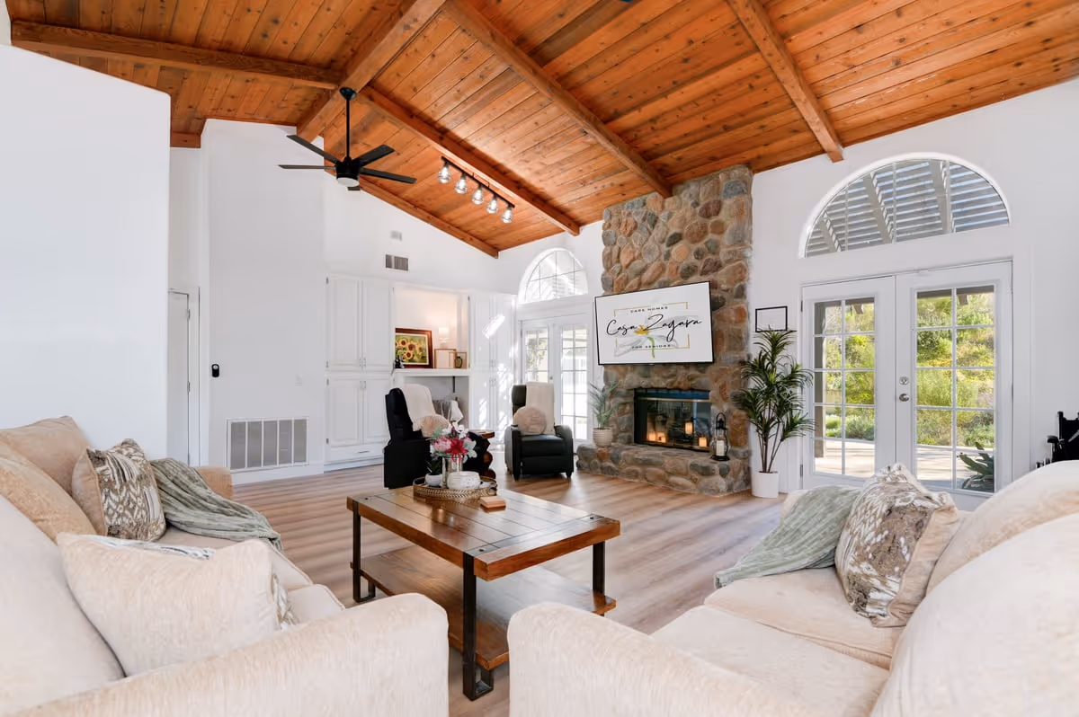 A spacious living room with a high wooden ceiling and exposed beams. The room features a stone fireplace with a mounted TV displaying the text 'Casa Zagara Inc.' Two comfortable armchairs are placed near the fireplace, and a wooden coffee table is centered between two beige sofas with decorative pillows and throws. Large windows and glass doors allow natural light to fill the room, and a potted plant adds greenery near the fireplace.