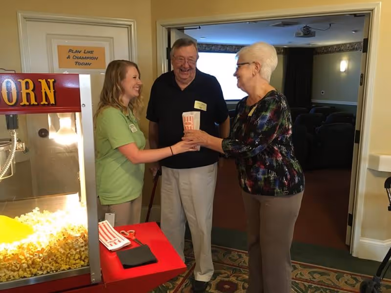 A young woman in a green shirt hands a popcorn container to an elderly woman with white hair and glasses, while an elderly man with a cane stands between them smiling. They are indoors near a popcorn machine, with a room containing theater-style seating visible in the background.