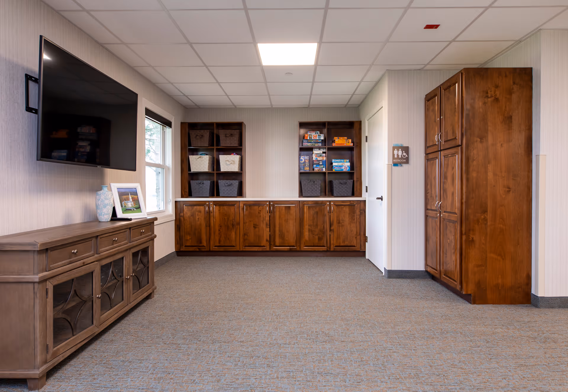 Bright common room with a wall-mounted TV, wooden cabinets and shelves holding baskets and games, and a closed door.