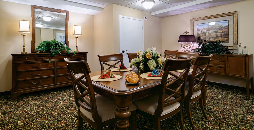 A dining room with a wooden table set for six, featuring floral centerpieces and folded napkins on placemats. The room has patterned carpet, two wooden sideboards with lamps and plants, a large mirror, and a framed painting on the wall.