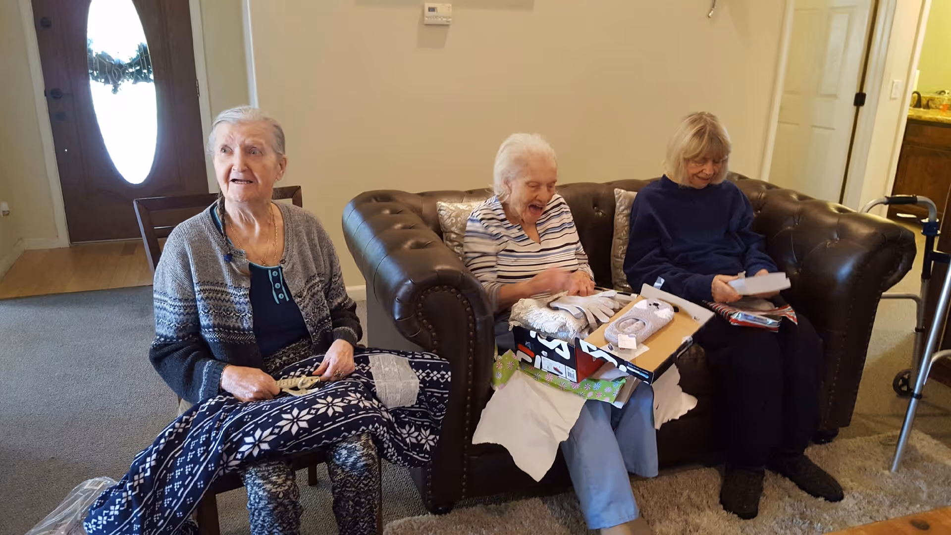 Three elderly women sitting in a living room of an assisted living facility. One woman is seated on a chair holding a patterned blanket, while the other two women are sitting on a brown leather couch, opening gifts and smiling. The room has a carpeted floor, a wooden front door with an oval window, and a walker is visible near the couch.