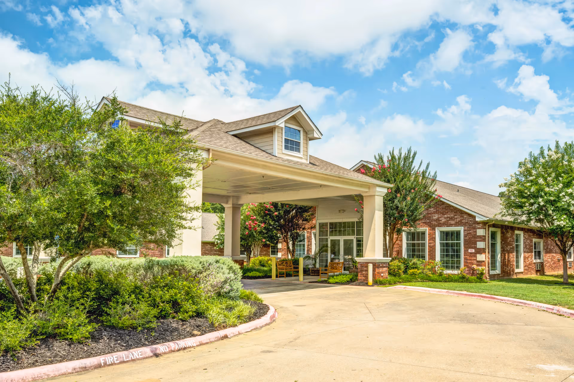 Front exterior view of Dogwood Trails Assisted Living and Memory Care facility showing a covered entrance with seating, surrounded by green bushes and trees under a partly cloudy sky.