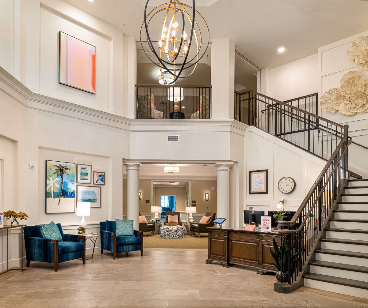 Spacious and elegant senior living facility lobby with high ceilings, a large modern chandelier, a wooden reception desk, a staircase with black railings, and comfortable seating areas with blue armchairs and decorative artwork on the walls.
