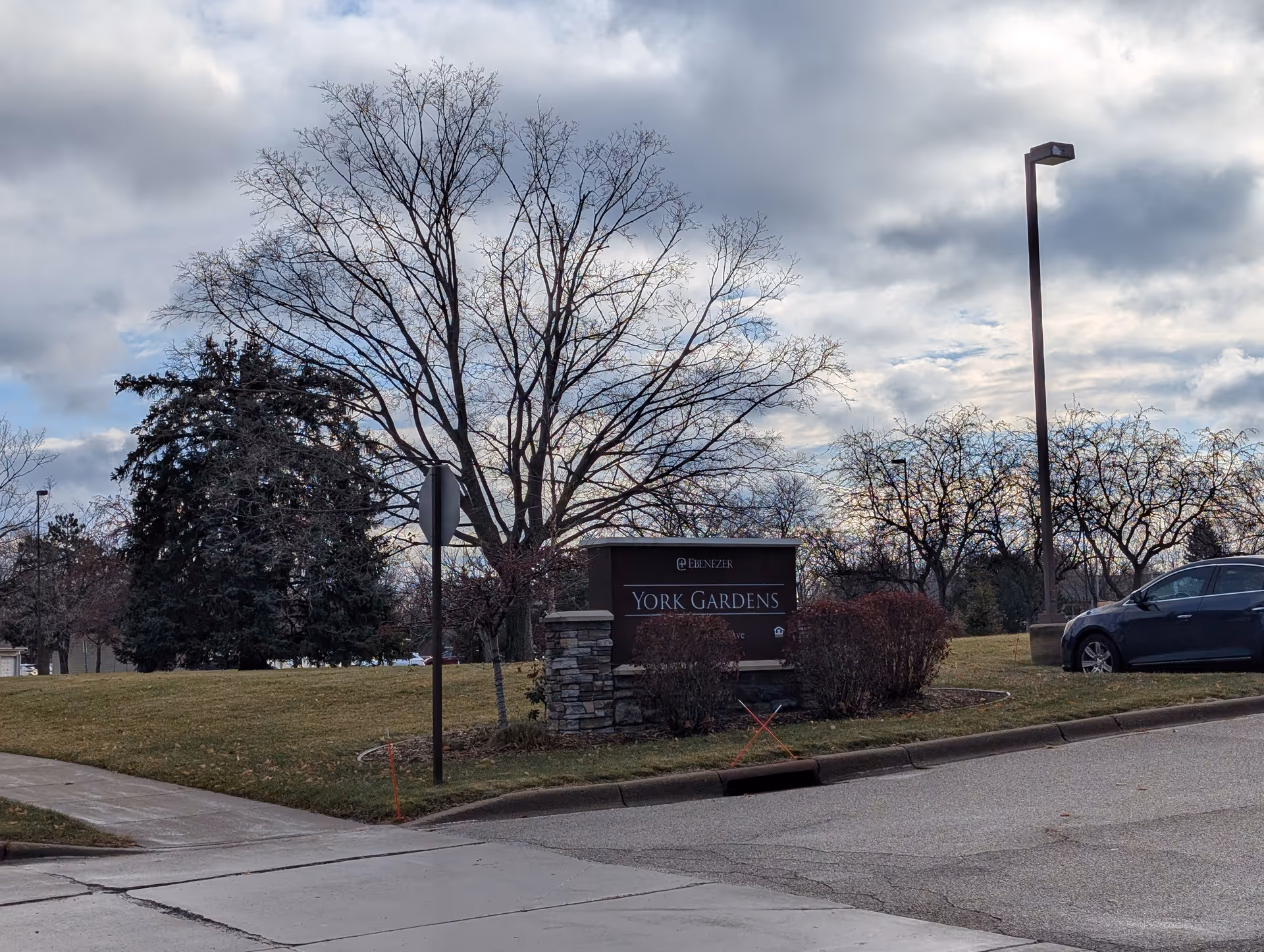 Entrance sign reading 'York Gardens' on a grassy corner with leafless trees, a streetlight and a parked car under a cloudy sky.