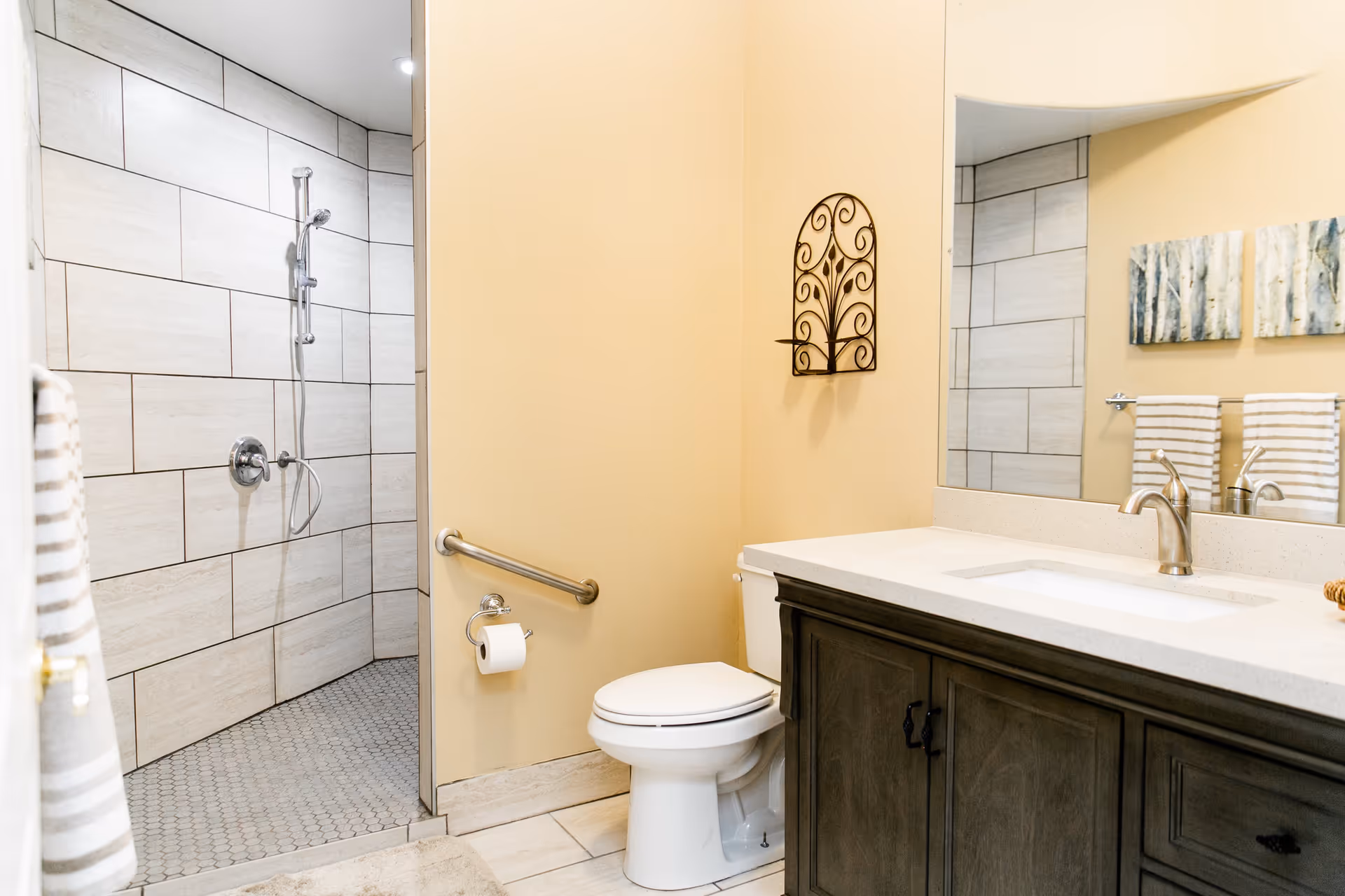 Bright bathroom featuring a tiled walk-in shower, toilet with a grab bar, and a vanity with sink and mirror.