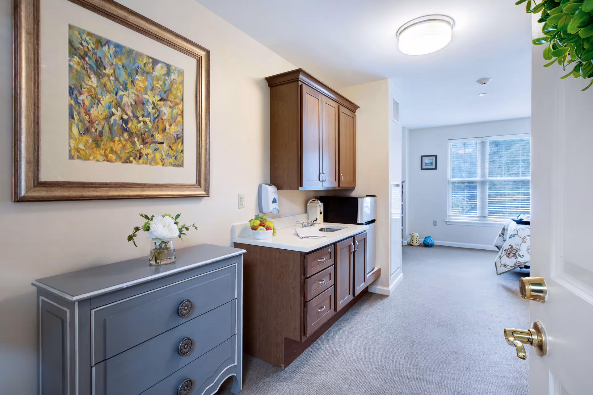 View into a bright senior living suite with a small kitchenette and wood cabinets, a gray dresser with flowers, and a bed by a window.