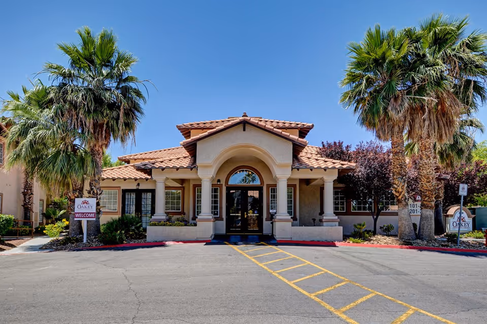 Front exterior view of Oakey Assisted Living facility with a tiled roof, arched entrance supported by columns, palm trees on both sides, and clear blue sky.