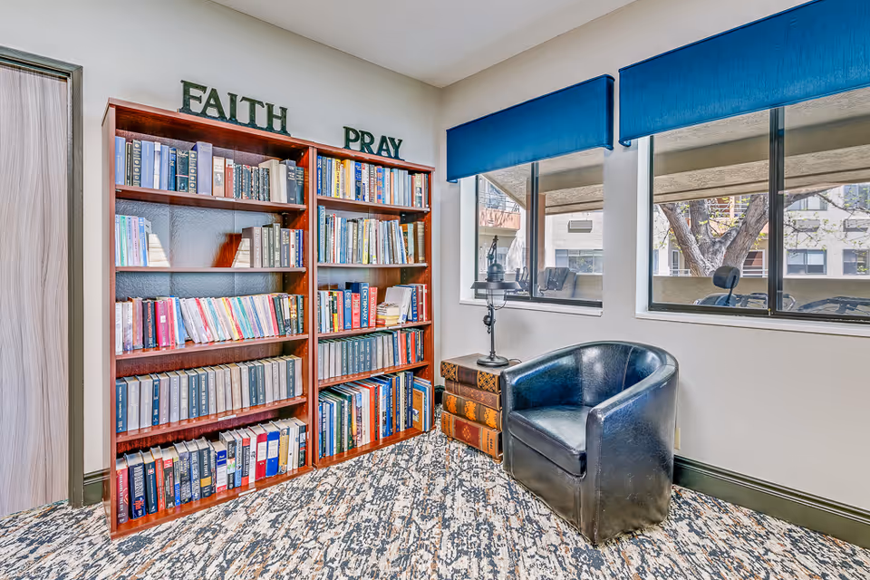 A cozy reading nook in a senior living facility featuring two wooden bookshelves filled with books. On top of the bookshelves are decorative letters spelling 'FAITH' and 'PRAY'. Next to the shelves is a small side table designed to look like stacked books with a candle holder on top. A black leather armchair is positioned nearby, and two windows with blue valances let in natural light, showing an outdoor view with trees and buildings.