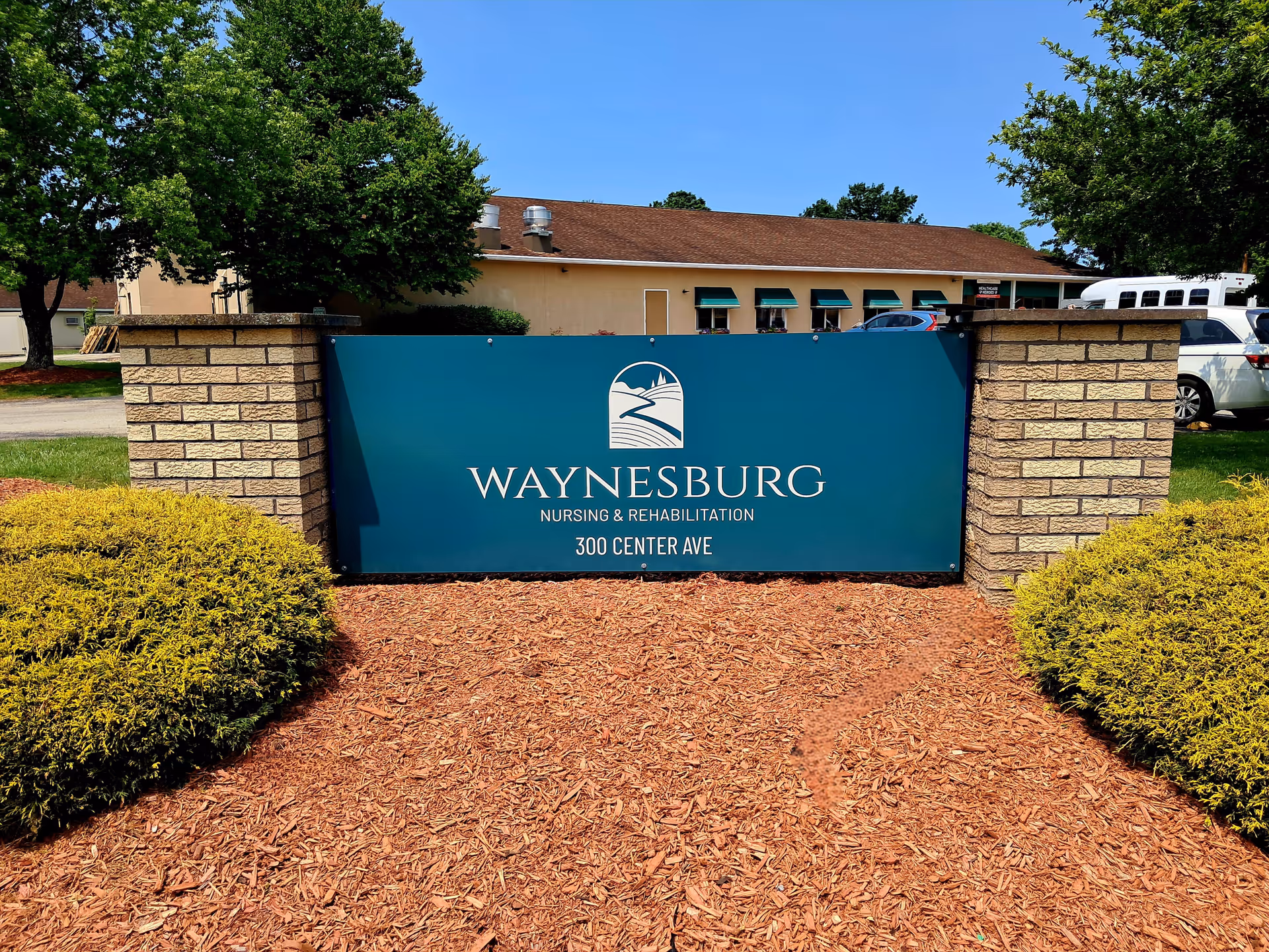 Outdoor view of the entrance sign for Waynesburg Nursing & Rehabilitation located at 300 Center Ave, surrounded by landscaped bushes and trees with a building and parked vehicles in the background under a clear blue sky.