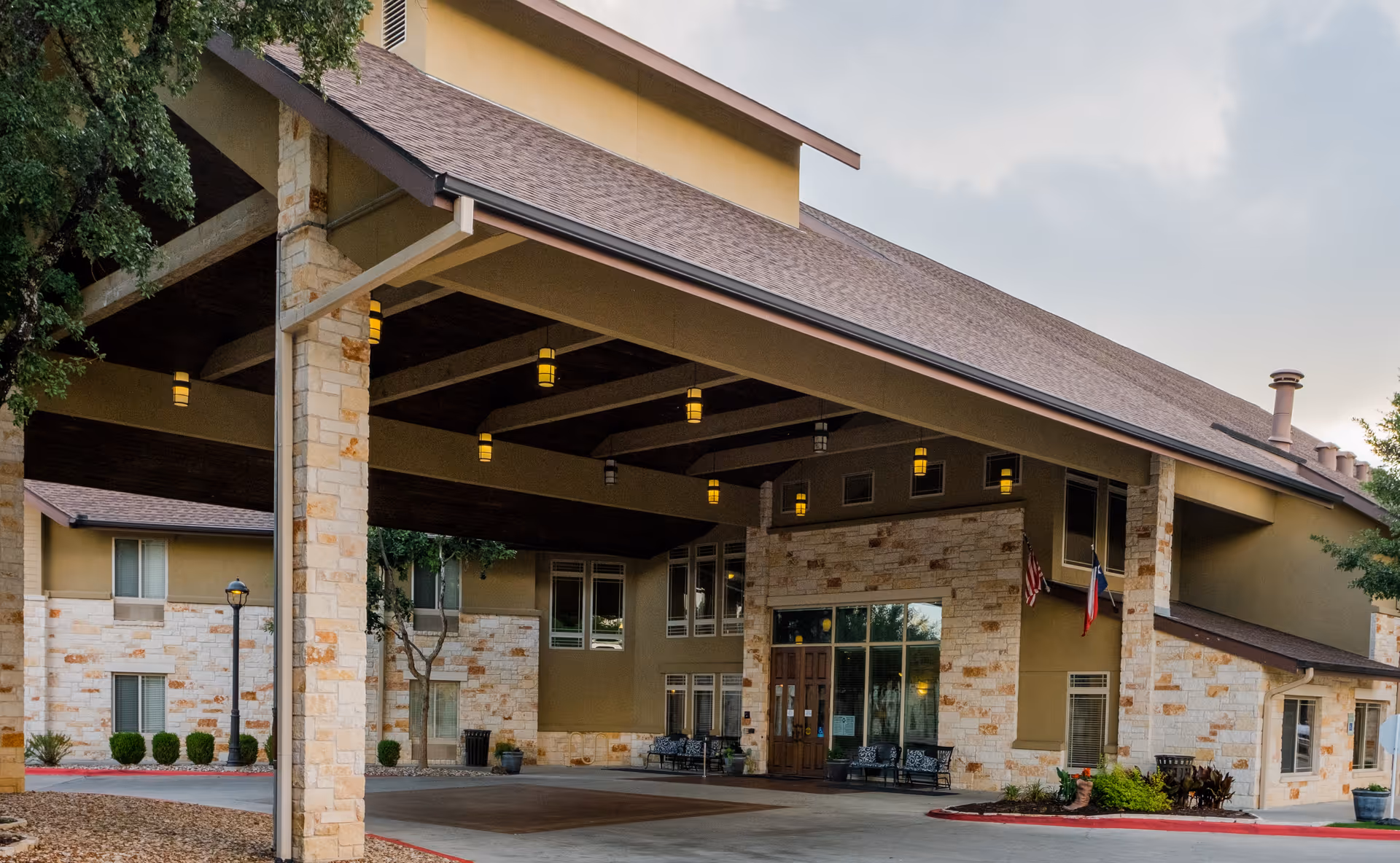 Entrance of The Pavilion at Great Hills facility showing a covered drop-off area with stone pillars, hanging lights, benches, and flags near the main door under a large roof.
