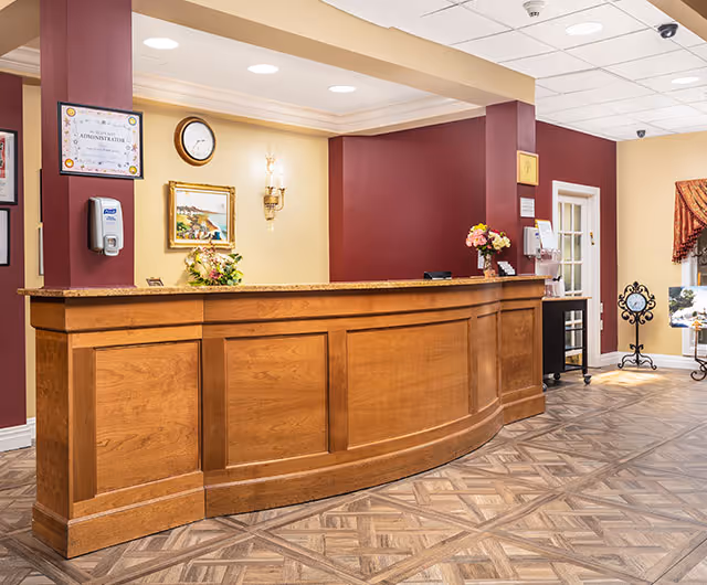 Reception area with a curved wooden front desk, maroon and beige walls, a clock, framed artwork, flowers on the counter, and a hand sanitizer dispenser mounted on a pillar.
