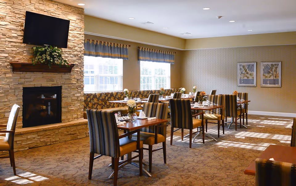 A dining room with several wooden tables and striped cushioned chairs arranged neatly. The room features a stone fireplace with a mounted flat-screen TV above it and a plant on the mantel. Large windows with valances allow natural light to fill the space. The walls are decorated with framed botanical artwork.