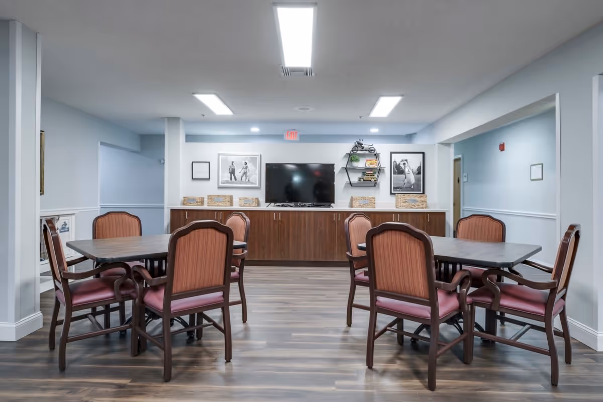 A common area with two rectangular tables surrounded by wooden chairs with red cushions. The room has light blue walls, a wooden floor, and a long cabinet against the back wall with a flat-screen TV mounted above it. There are framed black and white photos and decorative shelves on the wall behind the TV. The ceiling has bright rectangular lights.