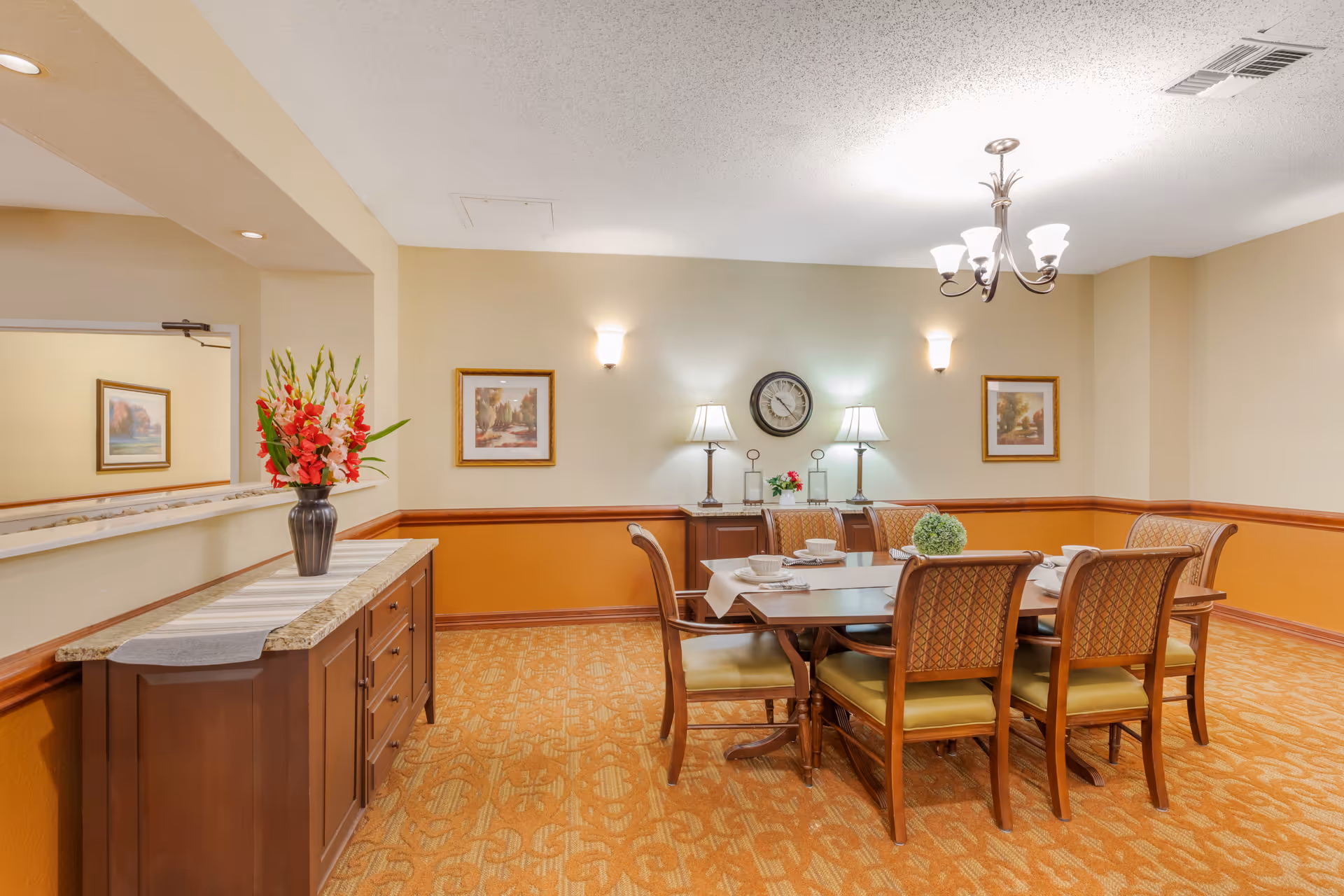 Well-lit dining room with a wooden table and six upholstered chairs, a sideboard with a vase of flowers, wall clock and framed art.