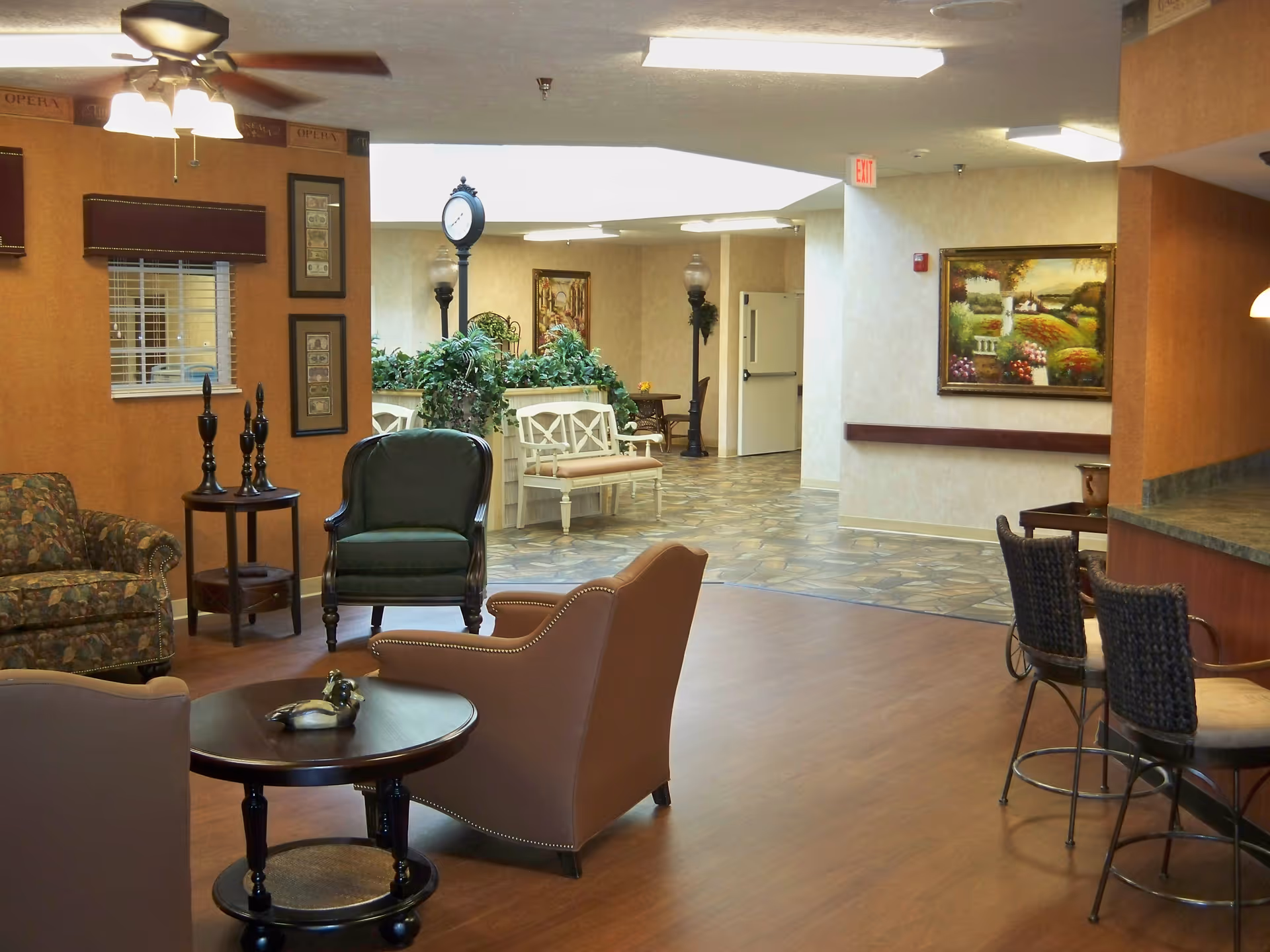 A cozy seating area in a retirement community with various chairs and a round wooden table. The room features warm-toned walls, a ceiling fan with lights, framed artwork, and decorative plants. In the background, there is a hallway with additional seating and a clock on a post.
