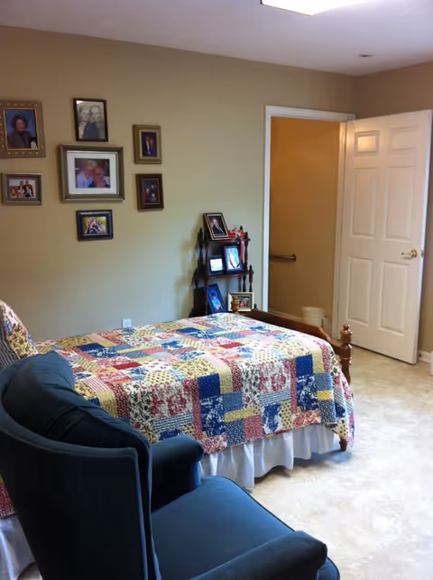 A bedroom with a single bed covered in a colorful patchwork quilt. There is a dark blue armchair in the foreground. The wall behind the bed has several framed photographs arranged in a gallery style. A small wooden shelf next to the bed holds more framed photos and decorative items. The door to the room is open, revealing a hallway with a grab bar on the wall.