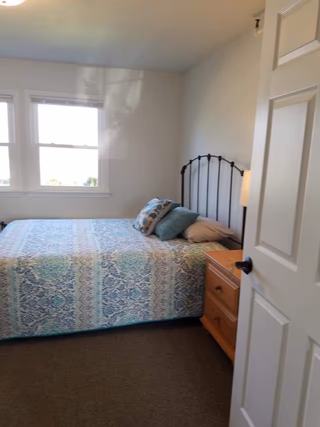 Small bedroom with a patterned bedspread, iron headboard, pillows, wooden nightstand and two windows.