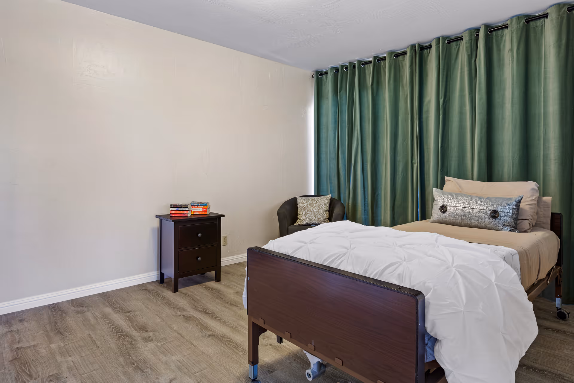 A simple bedroom with a single bed featuring a white comforter and beige pillows, a dark wooden nightstand with books on top, a dark armchair with a decorative pillow, and green curtains covering the window. The room has light-colored walls and wood flooring.