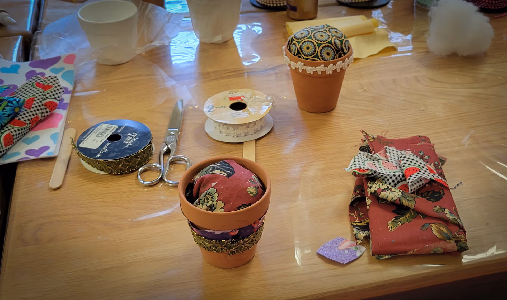 A wooden table with various craft supplies including two small decorated terracotta pots, fabric pieces, scissors, ribbon spools, a wooden stick, and a white cup. The table is covered with a clear plastic sheet.