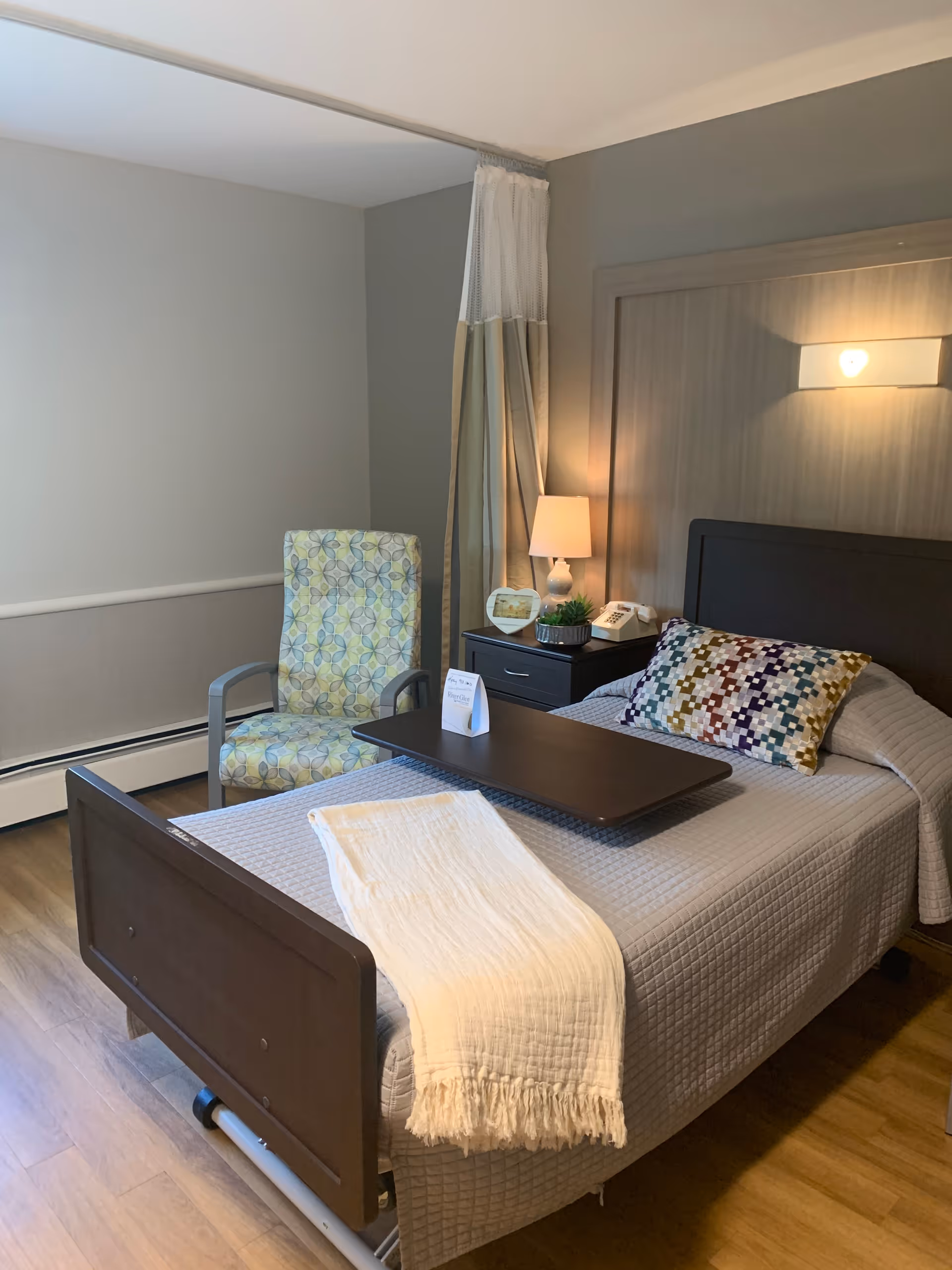 A neatly made single bed with a gray quilted bedspread and a colorful patterned pillow in a room. A white throw blanket is draped over the foot of the bed. There is a dark wooden bedside table with a lamp, a telephone, a small plant, and a photo frame. Next to the bed is a cushioned armchair with a geometric pattern. The room has light-colored walls and wood flooring.