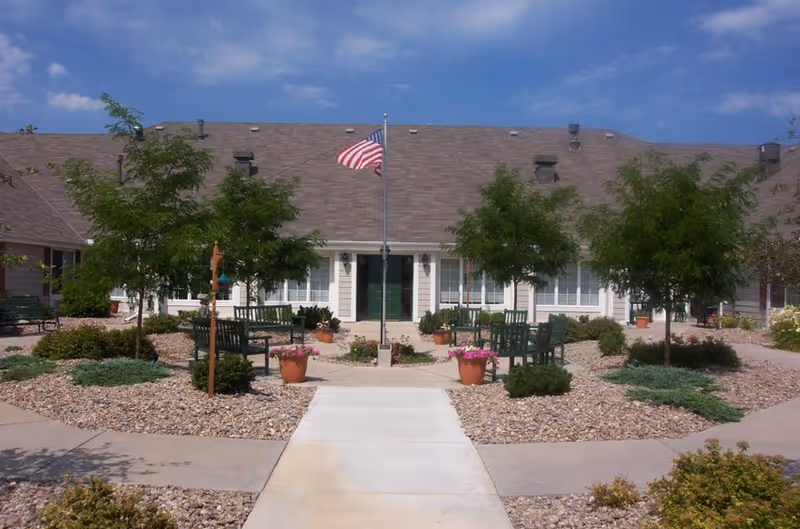 Outdoor courtyard area of a senior living facility with a concrete walkway leading to a building entrance. The courtyard has several green benches, potted plants with flowers, small trees, and an American flag on a flagpole in the center. The building has a brown shingled roof and white siding with multiple windows.