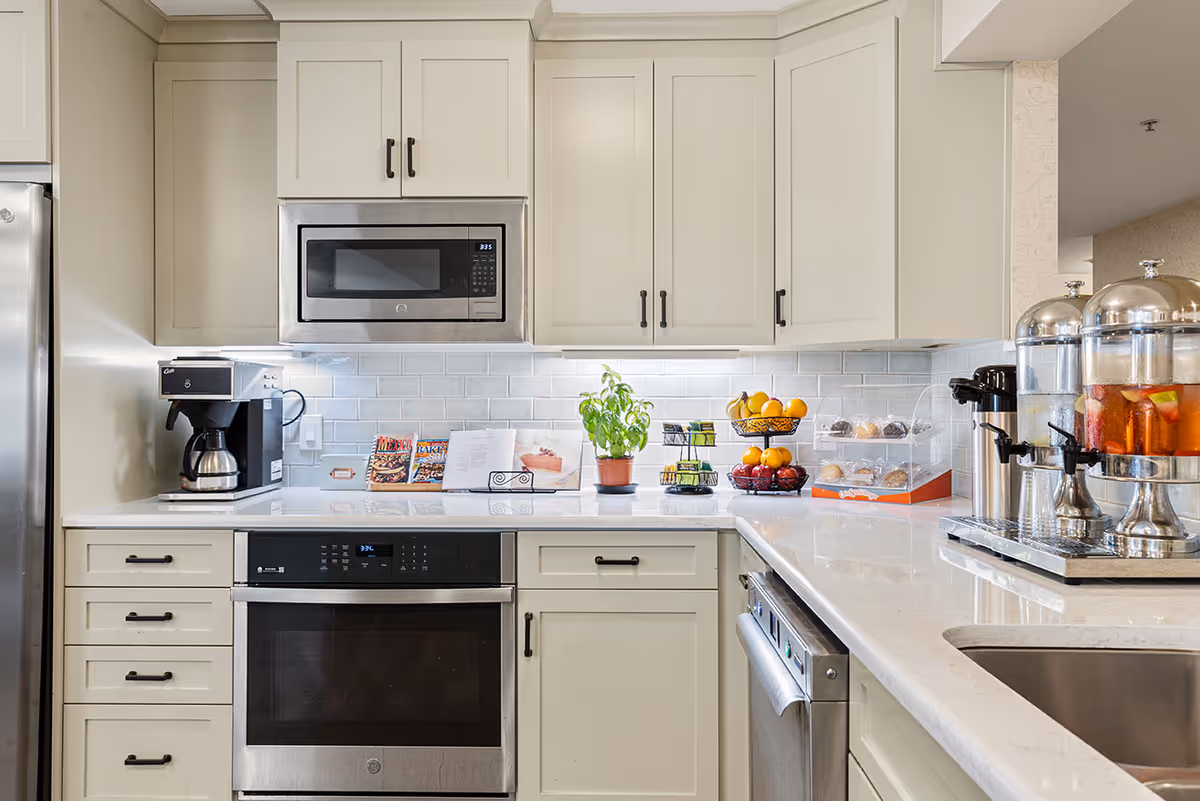 Modern kitchen with white cabinets, stainless steel oven and microwave, a coffee maker and beverage dispensers on the countertops, and bowls of fruit.