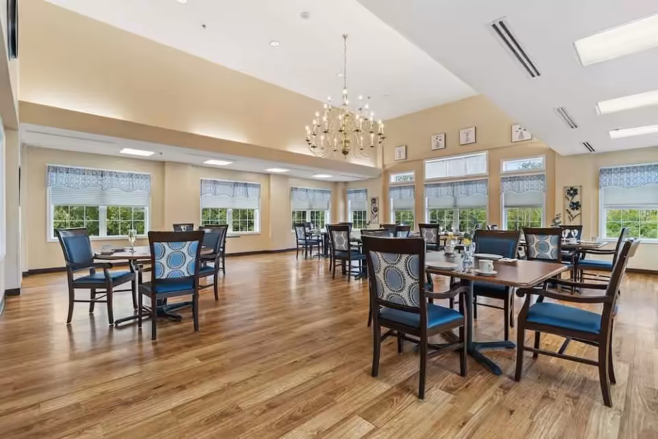 A spacious dining room with wooden floors, multiple tables and chairs with blue and patterned upholstery, large windows with white blinds and valances, and a chandelier hanging from the ceiling.