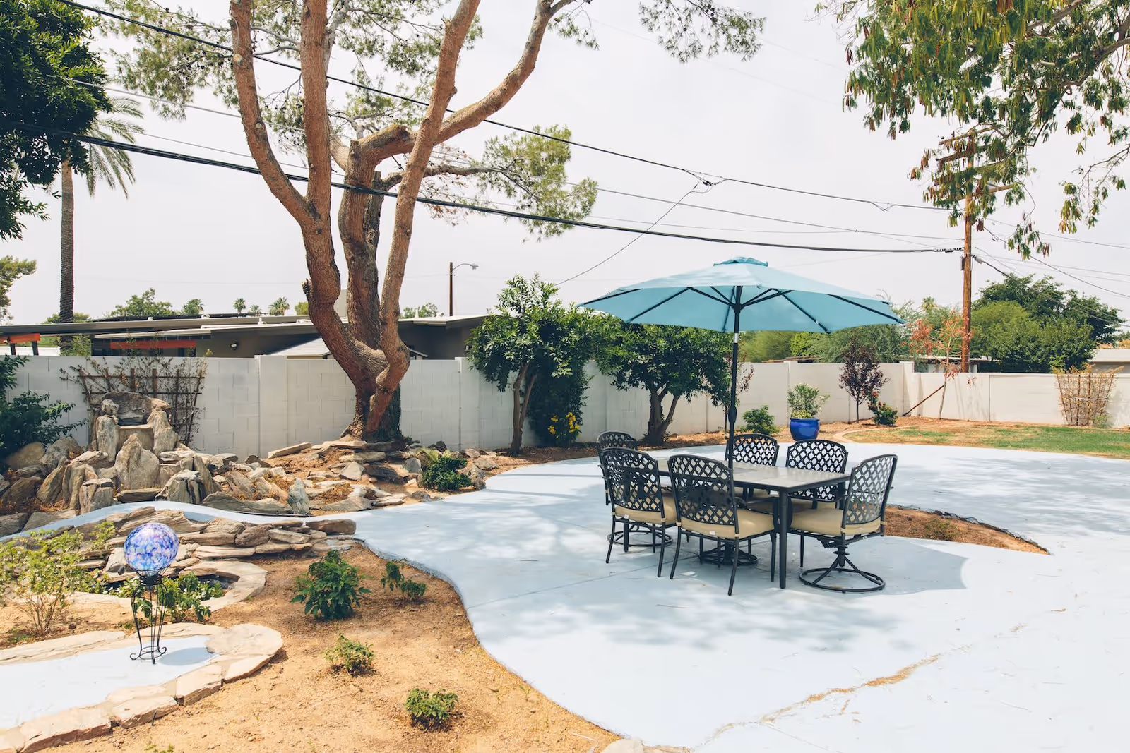 Outdoor patio area with a table and six chairs under a large blue umbrella. The patio is surrounded by a garden with small plants, trees, and rocks, enclosed by a white fence. Power lines and a building roof are visible in the background.