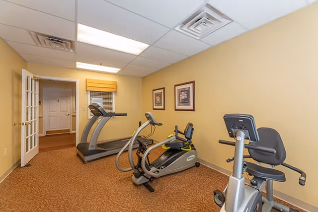 Small exercise room with a treadmill and two stationary/recumbent bikes against beige walls.