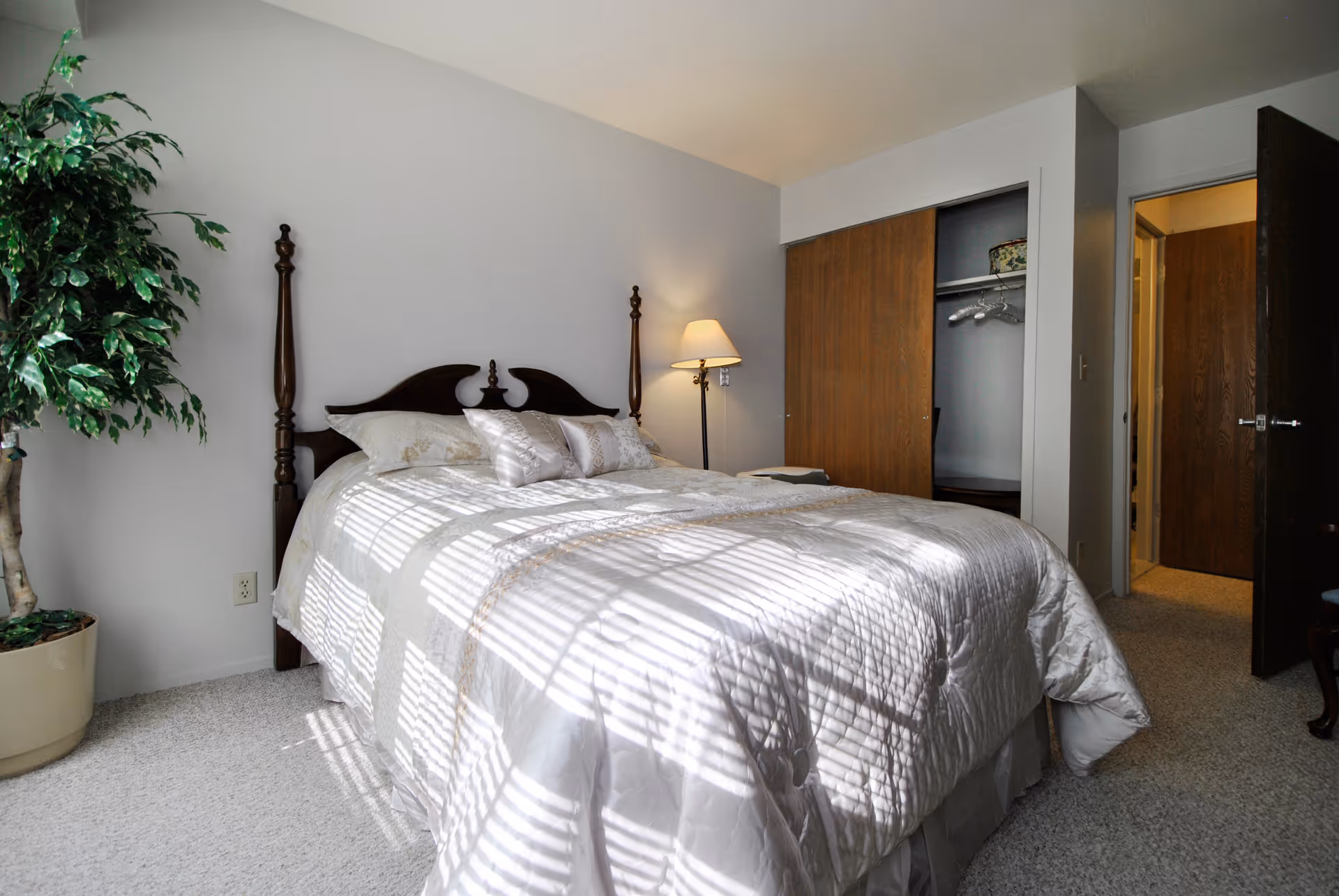 Sunlit bedroom with a made bed featuring a wooden headboard, bedside lamp, open closet, and potted plant.