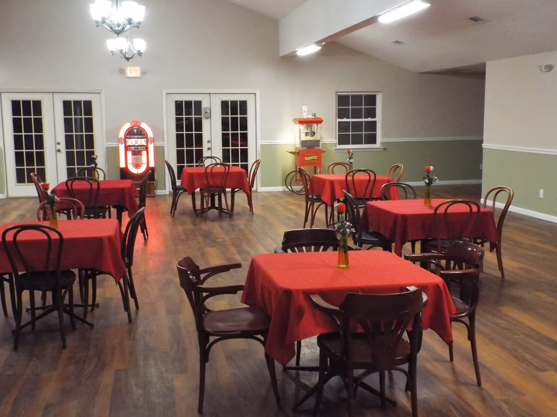 Interior view of a dining area with multiple tables covered in red tablecloths, each with a small vase holding a single red flower. The room has wooden flooring, light green and beige walls, and a ceiling light fixture. In the background, there is a jukebox and a popcorn machine near glass double doors and a window.