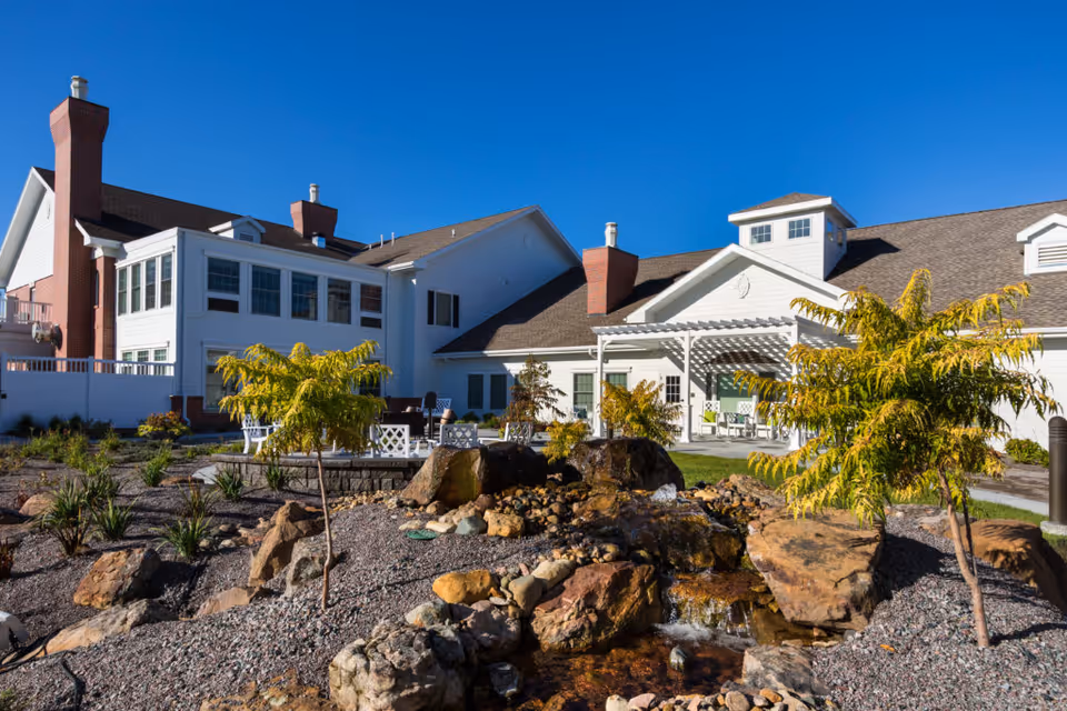 Front exterior of a white CountryHouse Residence building with a landscaped rock garden, small waterfall, and outdoor seating.