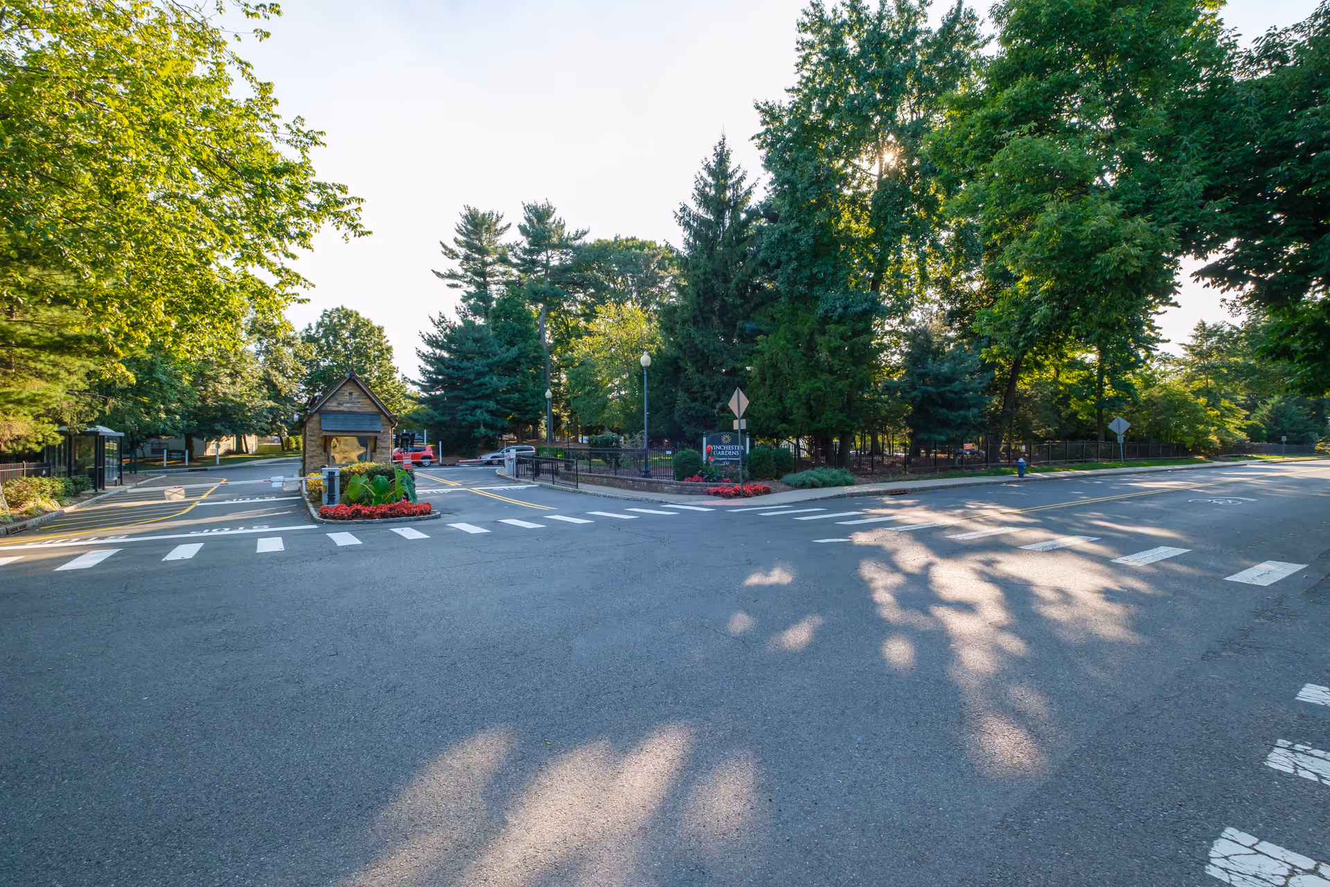 Entrance road to Winchester Gardens facility with a small guardhouse in the center, surrounded by trees and greenery on both sides. There are road markings and a sign indicating the facility name near the entrance.