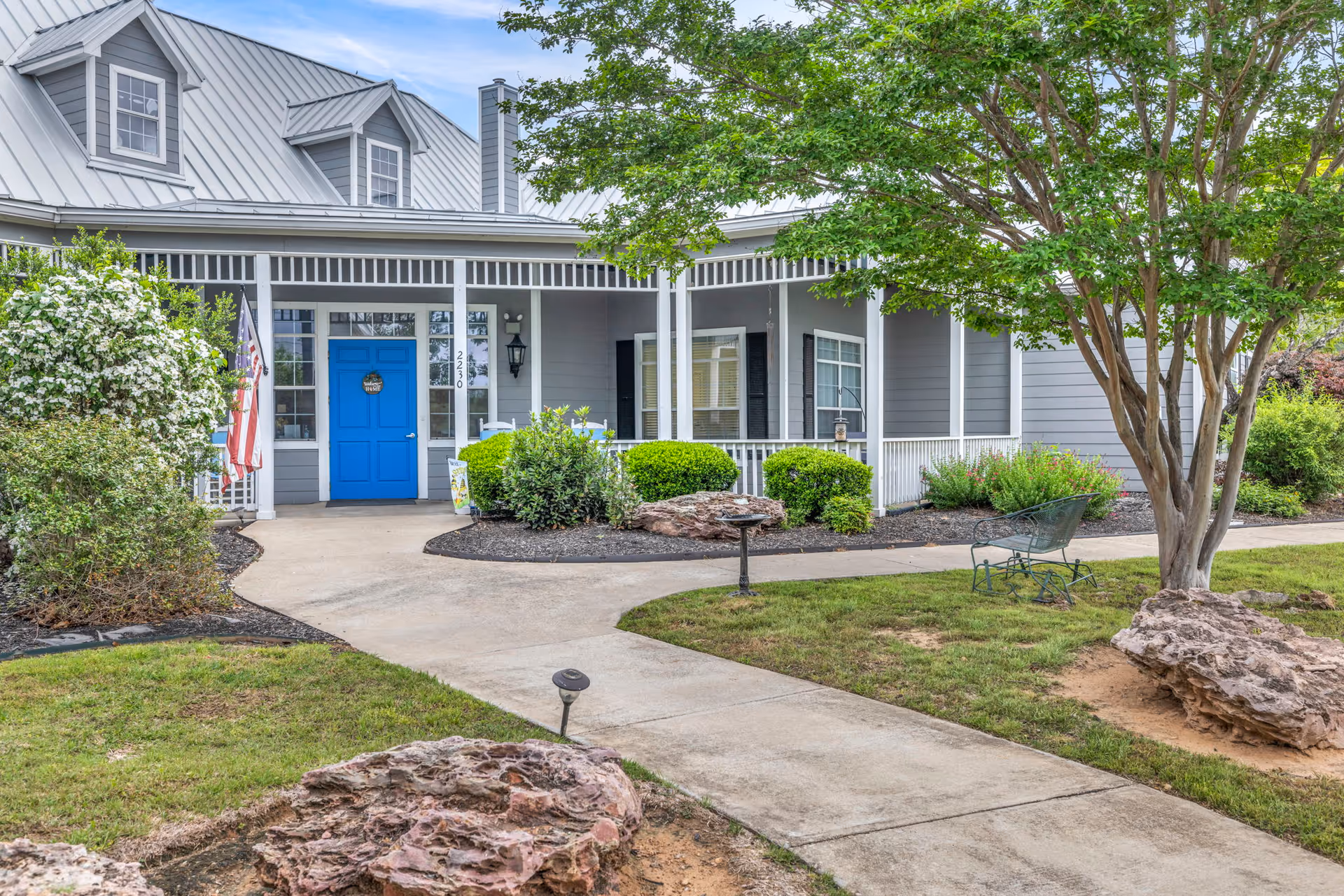 Front exterior of a gray, house-style building with a bright blue front door, wraparound porch, walkway, and landscaped greenery.