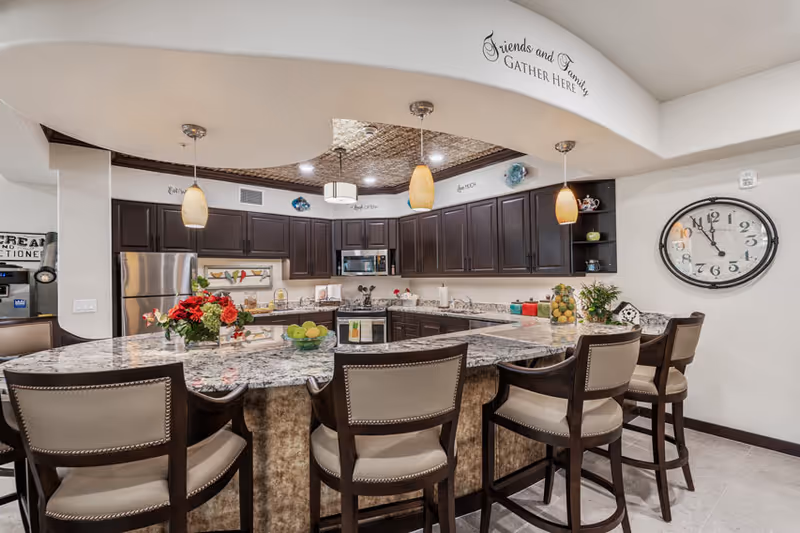 Modern kitchen area with a large granite countertop island surrounded by six cushioned bar stools. The kitchen features dark wood cabinets, stainless steel refrigerator, microwave, and stove. Three pendant lights hang above the island. A large wall clock is mounted on the right wall. Above the kitchen archway, there is a decorative sign that reads 'Friends and Family Gather Here'.