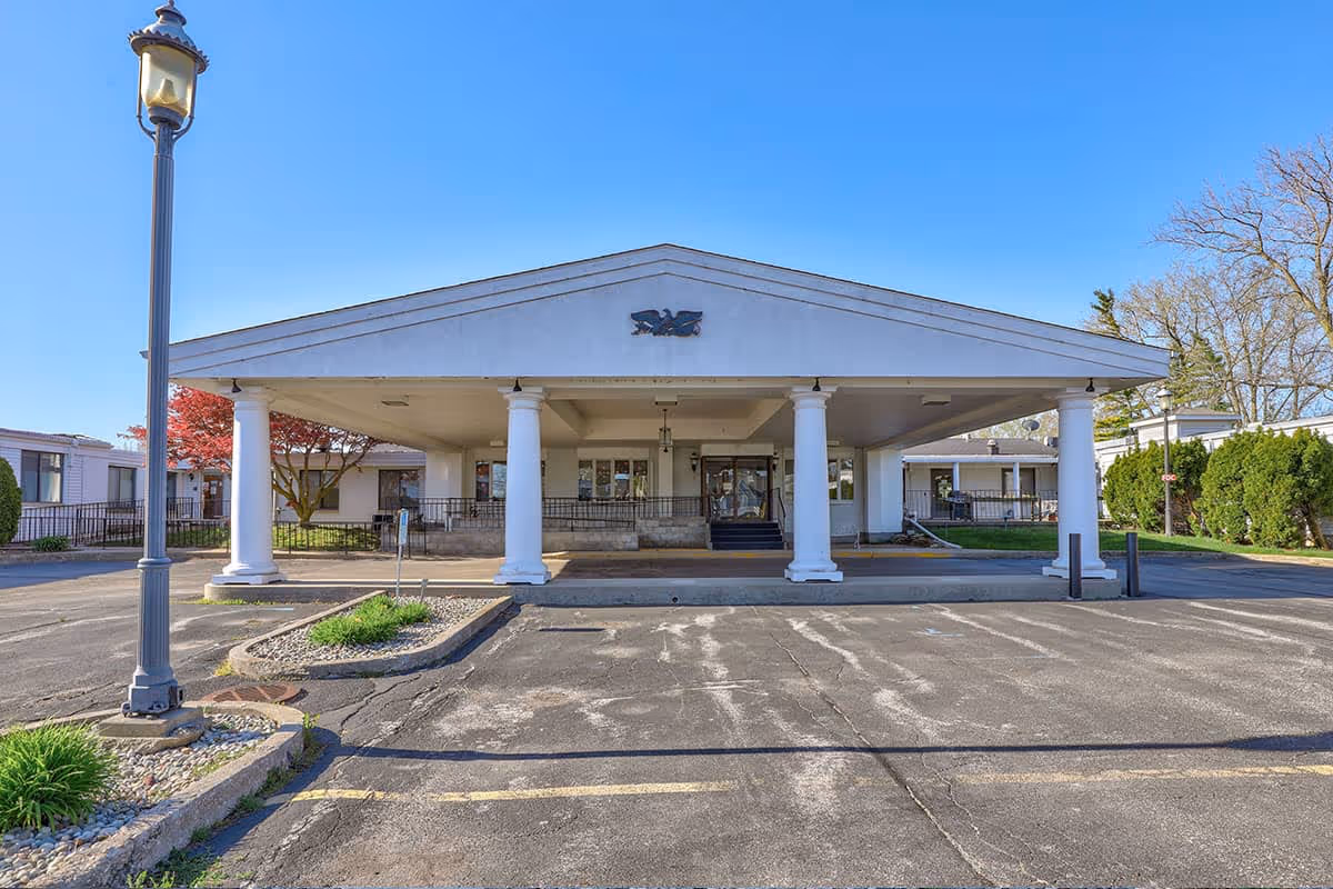 Front exterior view of Carriage House Nursing and Rehab building with a covered entrance supported by white columns, a lamp post on the left, and a clear blue sky.