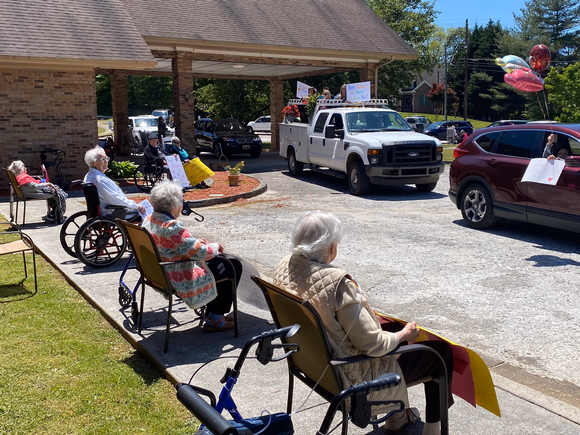 Several elderly individuals sitting outside a retirement facility, some in wheelchairs and others in chairs, watching a parade of vehicles passing by. The vehicles have people holding signs and balloons, and the scene is sunny with green trees and a brick building in the background.