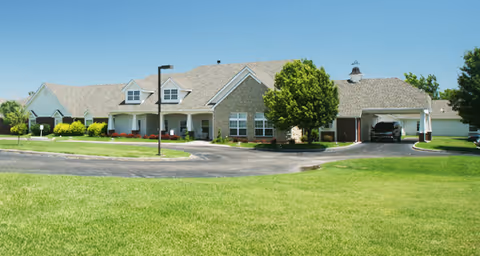 Exterior view of a single-story senior living facility building with a light-colored roof and stone facade, surrounded by green grass and trees under a clear blue sky.