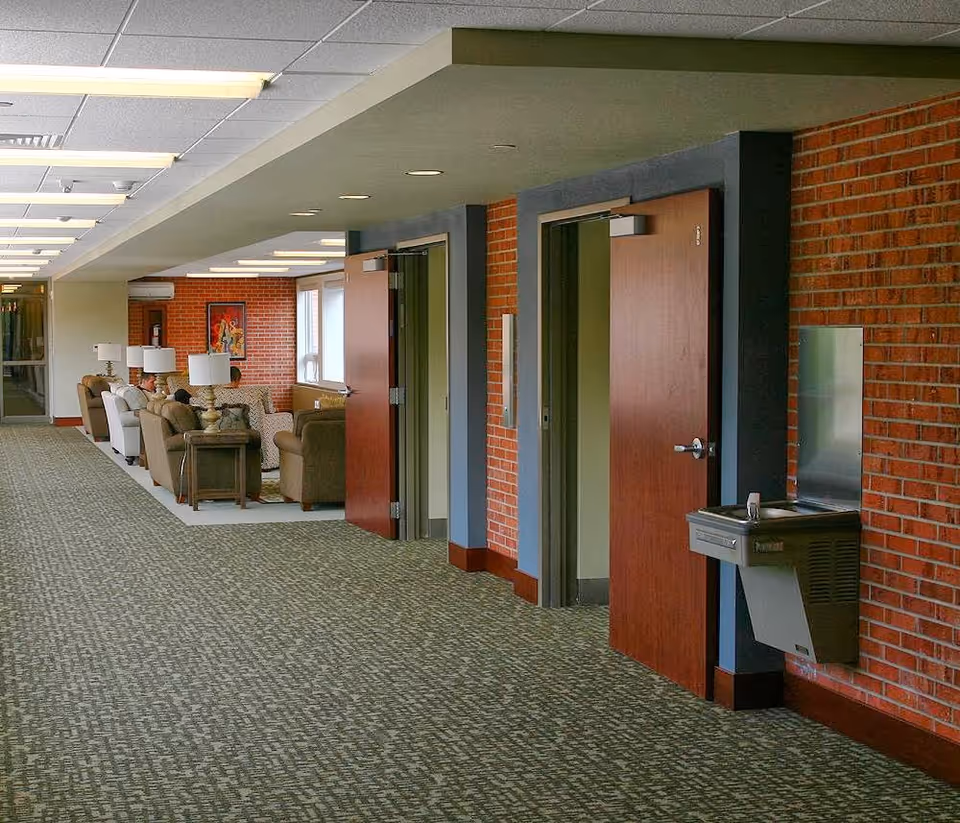 A long hallway with patterned carpet and brick walls on the right side. There are two open wooden doors leading to rooms, and a water fountain mounted on the wall. On the left side, there is a seating area with several armchairs, side tables, and lamps, where a few people are sitting. The ceiling has recessed lighting and fluorescent lights.