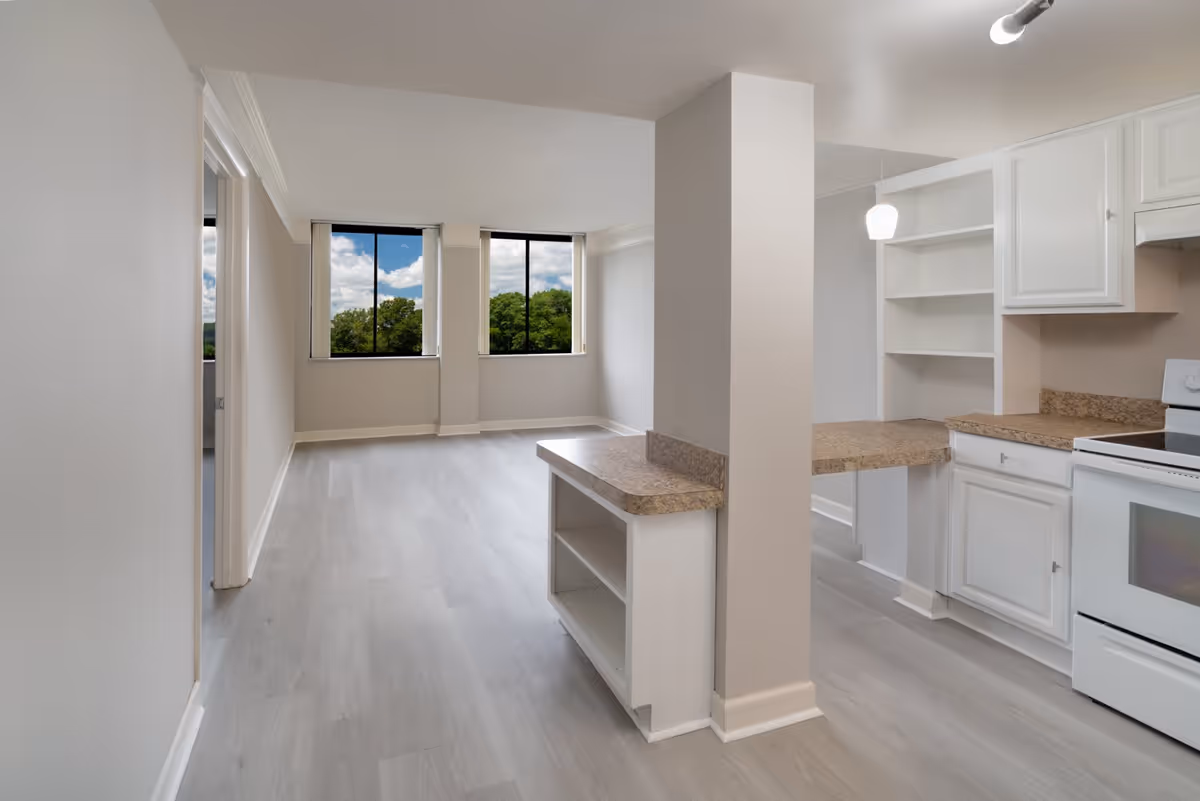 Open-plan apartment interior showing a kitchen with white cabinets and countertops opening into a bright living area with large windows and light wood flooring.