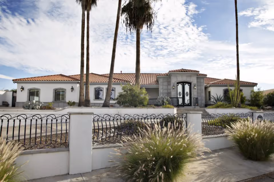 Front exterior of a single-story Mediterranean-style home with a tiled roof, iron gate, and palm trees in the landscaped front yard.