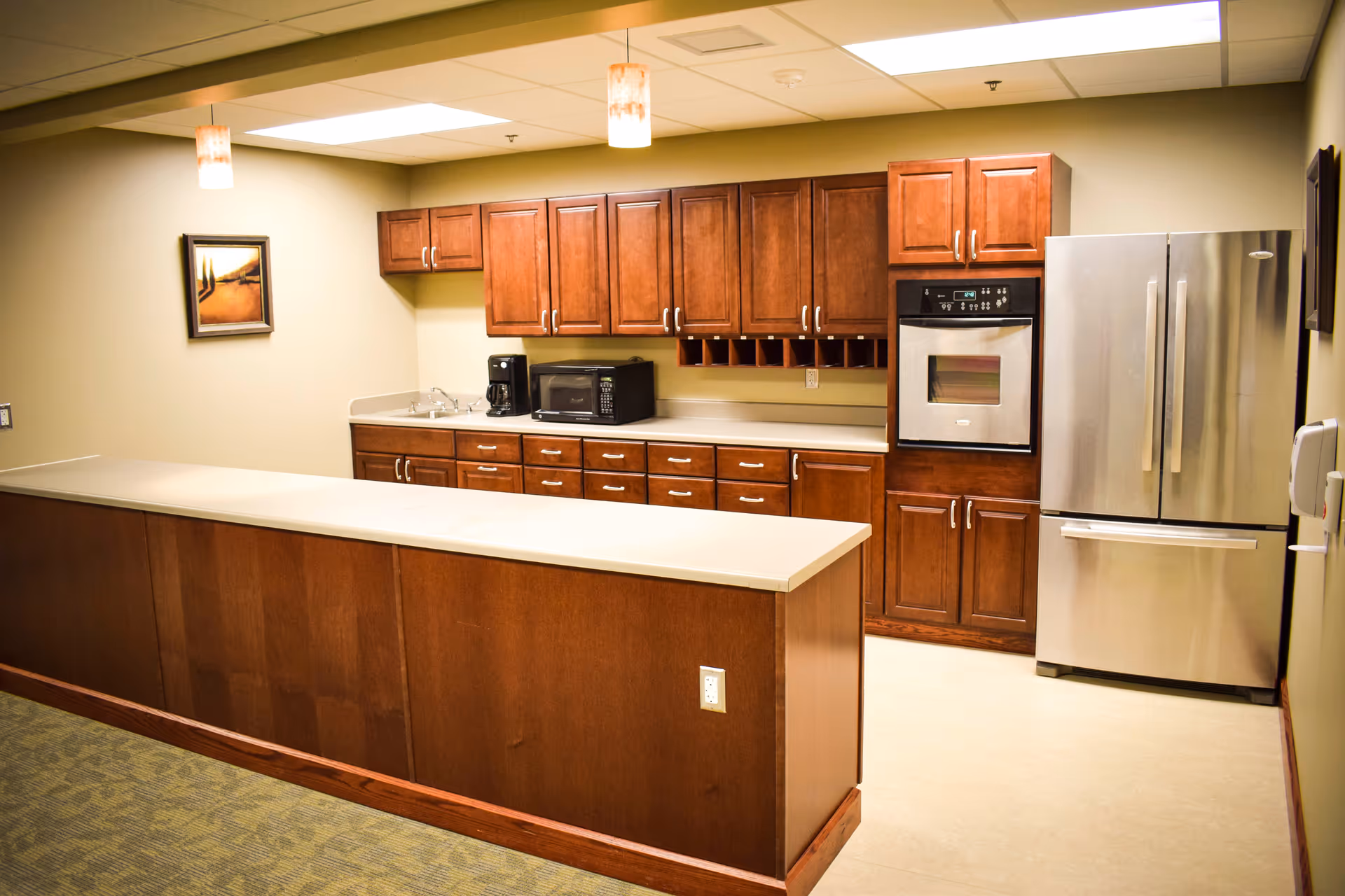 A kitchen area with wooden cabinets, a stainless steel refrigerator, built-in oven, microwave, coffee maker, and a sink. The room has a beige countertop and two pendant lights hanging from the ceiling. There is a framed painting on the wall and a carpeted floor outside the kitchen area.