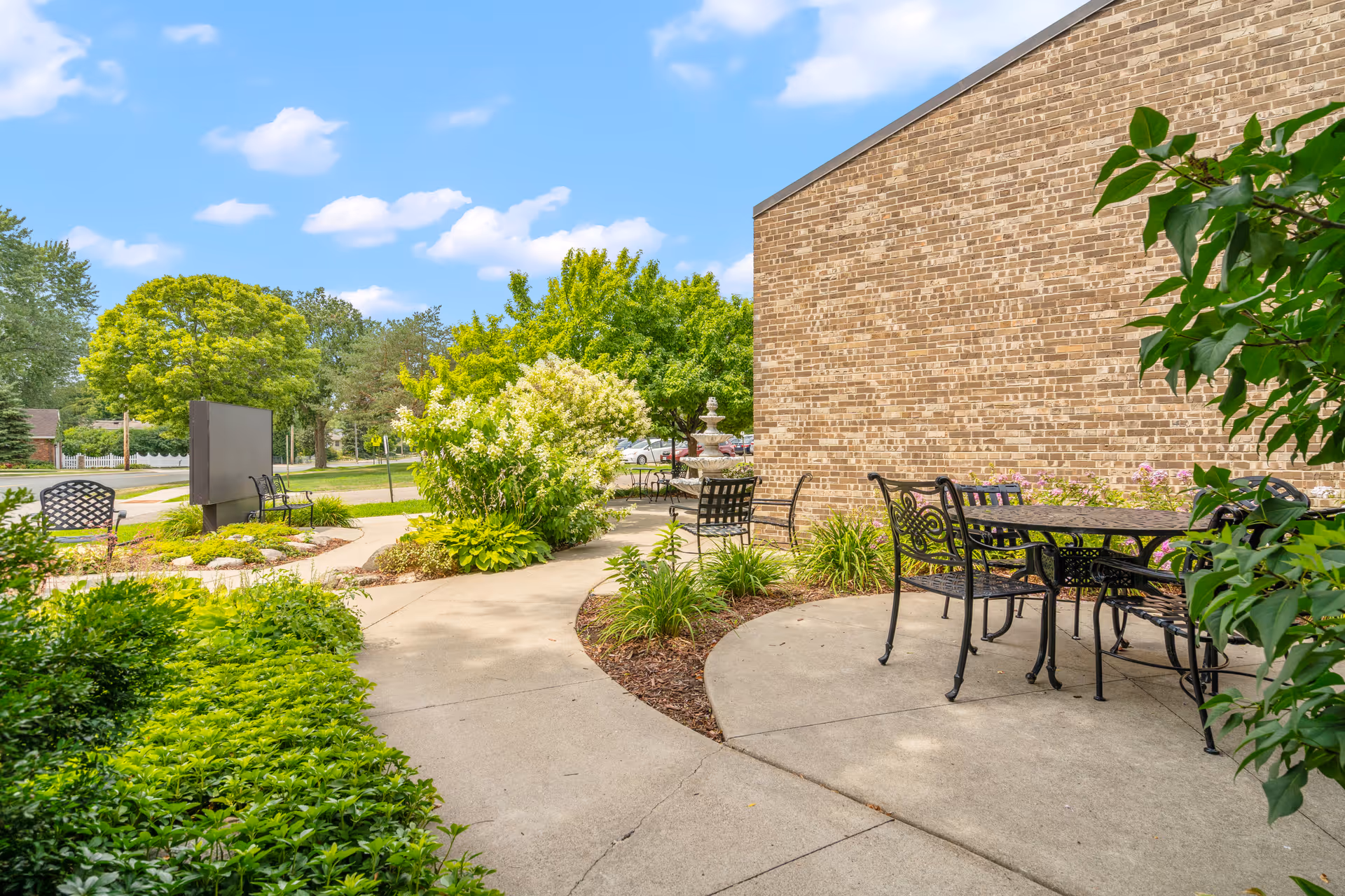 Outdoor patio area at Chapel View Health Care Center featuring black metal tables and chairs on a concrete surface, surrounded by green shrubs, trees, and a brick building wall under a blue sky with scattered clouds.