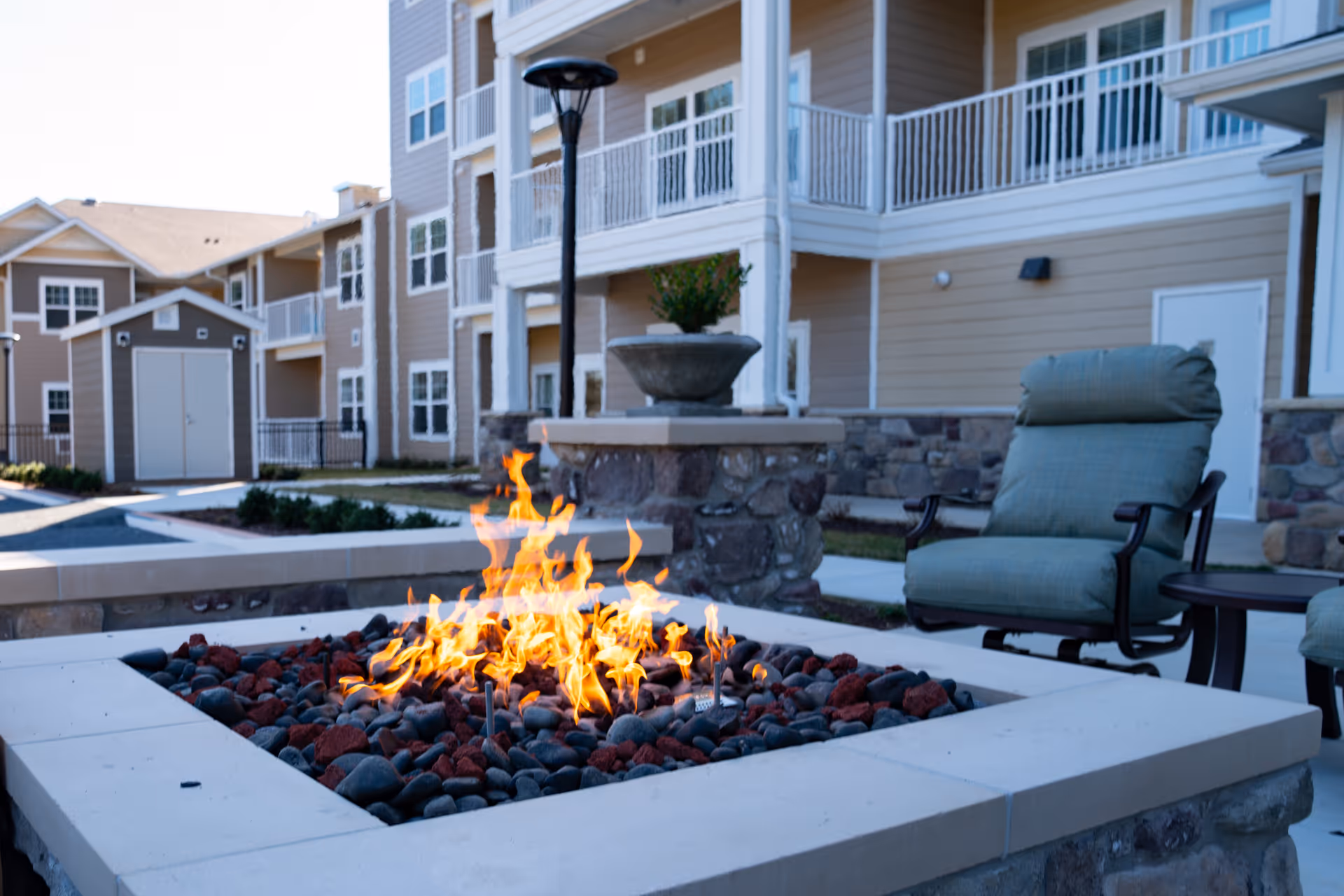 Outdoor seating area at a senior living facility with a stone fire pit burning surrounded by cushioned chairs. In the background, there are multi-story beige buildings with balconies and a small shed.
