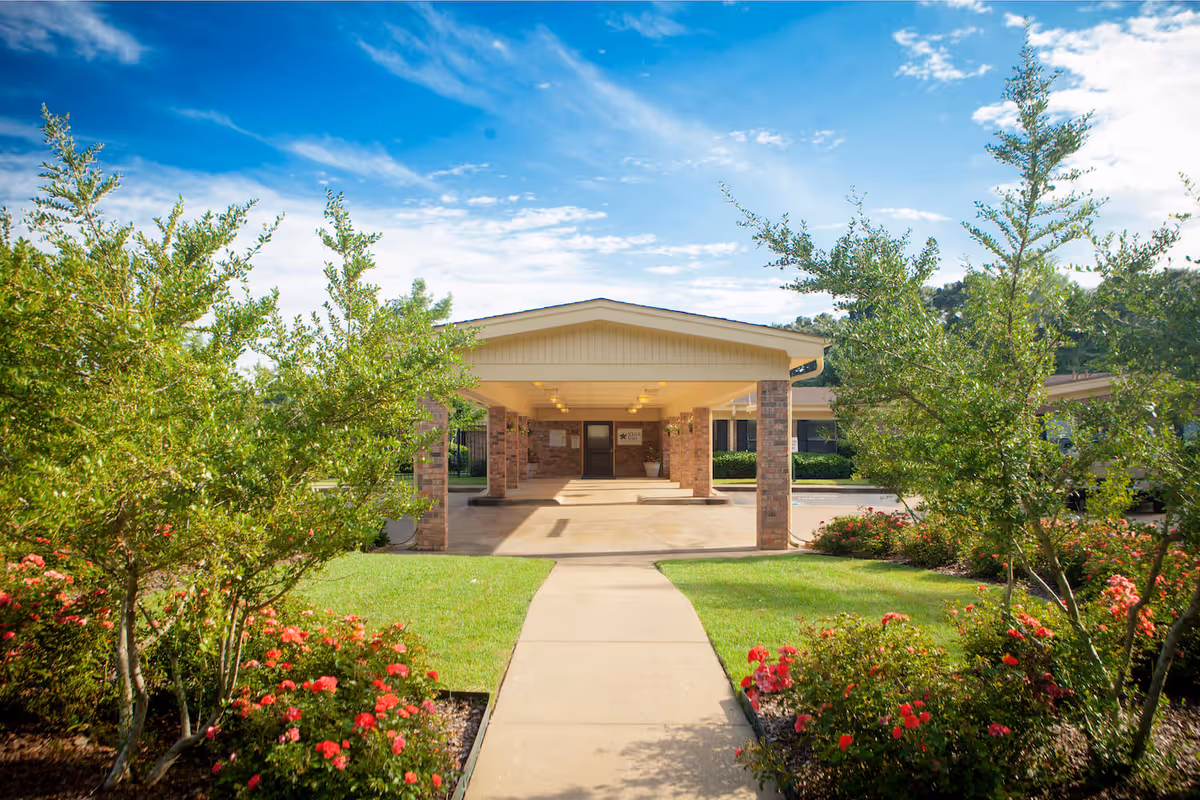 Front entrance of Briarcliff Health Center with a covered walkway supported by brick columns, surrounded by green grass, bushes, and flowering plants under a partly cloudy blue sky.