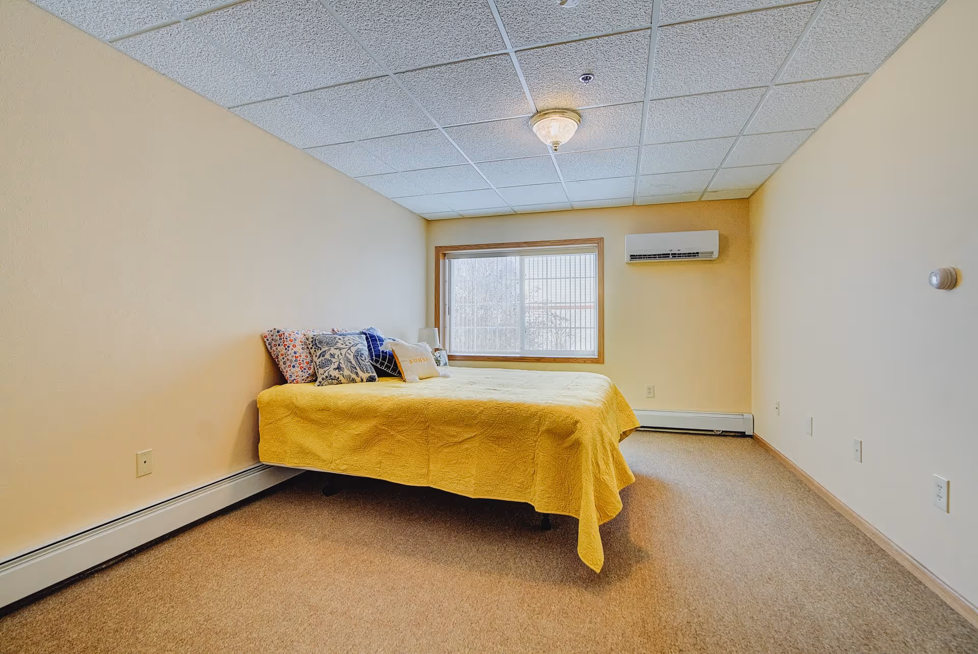 A simple bedroom with beige walls and carpeted floor. There is a bed with a yellow bedspread and several decorative pillows against the left wall. A large window with wooden trim is on the far wall, and a wall-mounted air conditioning unit is installed to the right of the window. The ceiling has white acoustic tiles with a central light fixture.