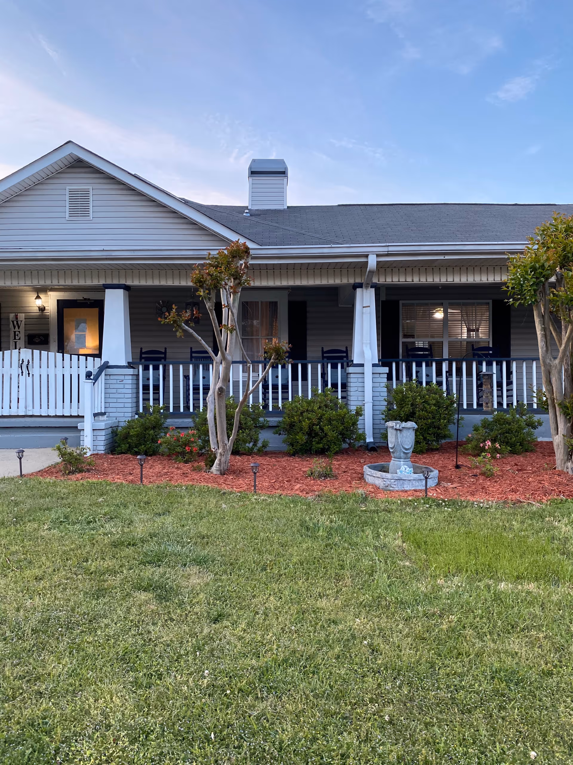 Front exterior view of a single-story senior living facility with a covered porch, white railing, rocking chairs, small trees, bushes, and a grassy lawn under a clear sky.