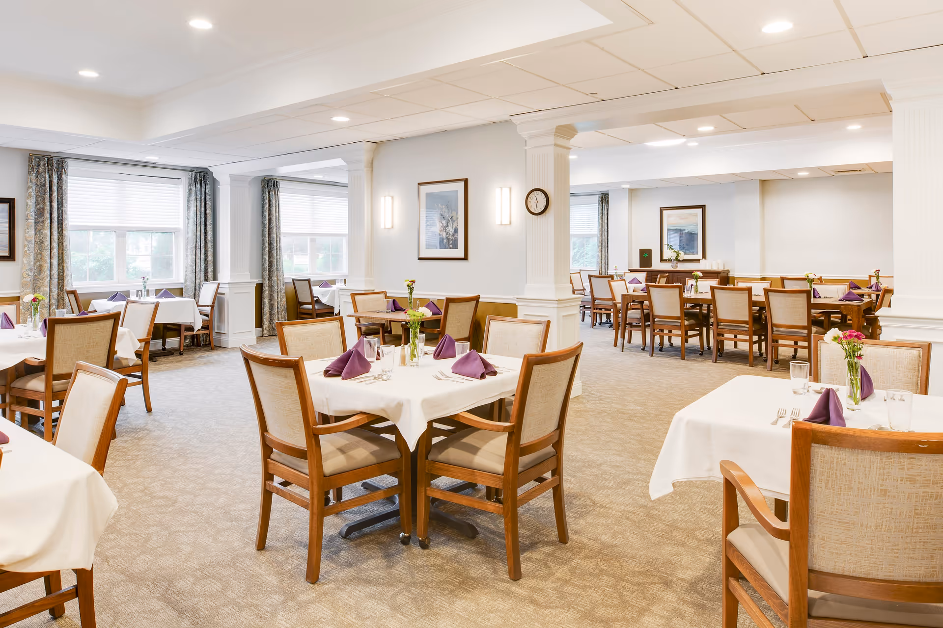 A bright and spacious dining room with multiple tables covered in white tablecloths, each set with purple folded napkins, glassware, and small flower vases. The room features large windows with curtains, beige carpet, and framed artwork on the walls.