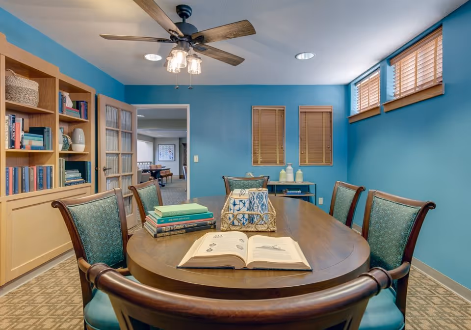 A cozy meeting or reading room with a wooden oval table surrounded by six chairs with teal patterned upholstery. On the table are several stacked books, an open book, and decorative items. The walls are painted teal, with two small windows with wooden blinds and three higher windows letting in natural light. A wooden bookshelf filled with books and decorative items is on the left, and a ceiling fan with lights hangs above the table. Through an open door, another room with seating is visible.